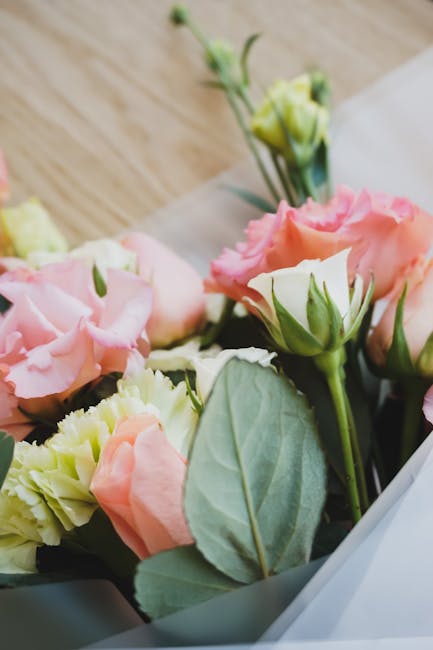 A close-up of a floral bouquet featuring soft pink and creamy white lisianthus flowers with delicate, ruffled petals. The arrangement includes lush green leaves with prominent veins, and some buds sti
