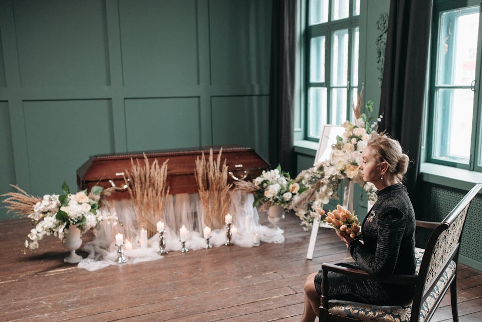A woman seated in a dark-patterned dress, holding a bouquet of soft peach and cream flowers, in a softly lit room with green walls and large windows. In front of her, a floral arrangement featuring wh